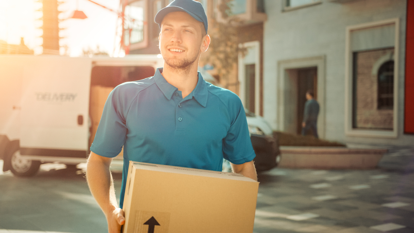 Portrait of Delivery Man Holds Cardboard Box Package Standing in Modern Stylish Business District with Delivery Van in Background. Smiling Courier On Way to Deliver Postal Parcel to Client. Sun Flare